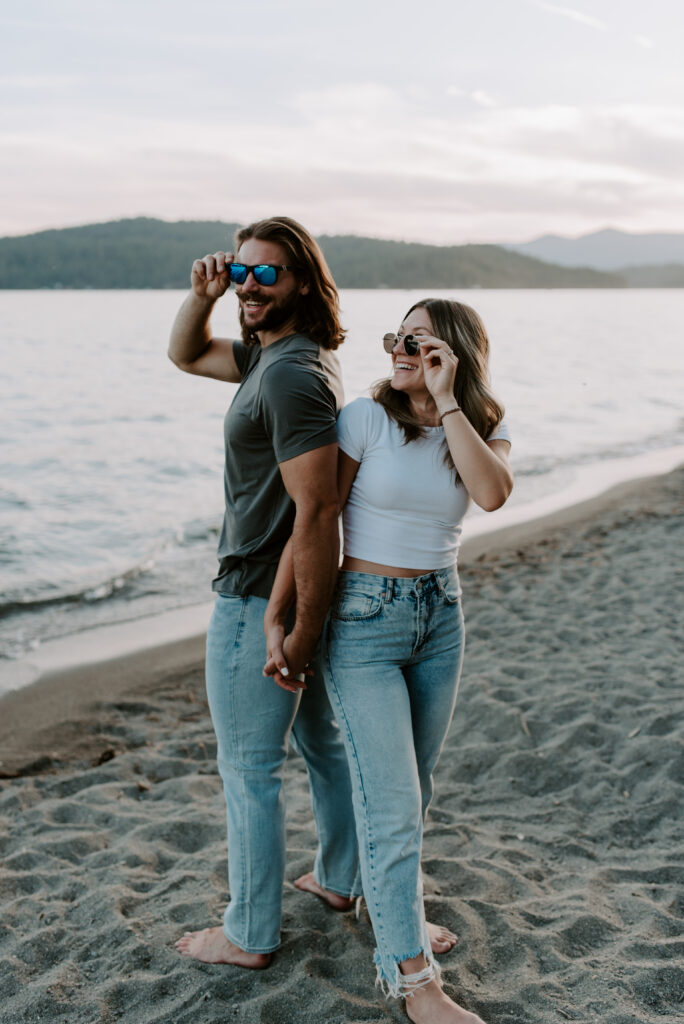 Couple with sunglasses holding hands and laughing on the beach at Lake Coeur D'Alene