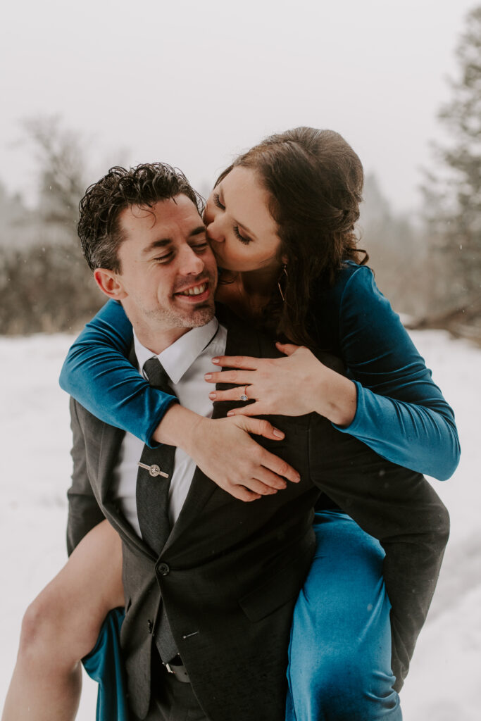 A future bride kisses her fiance on the cheek while doing a piggyback ride in the snow for their winter photos.