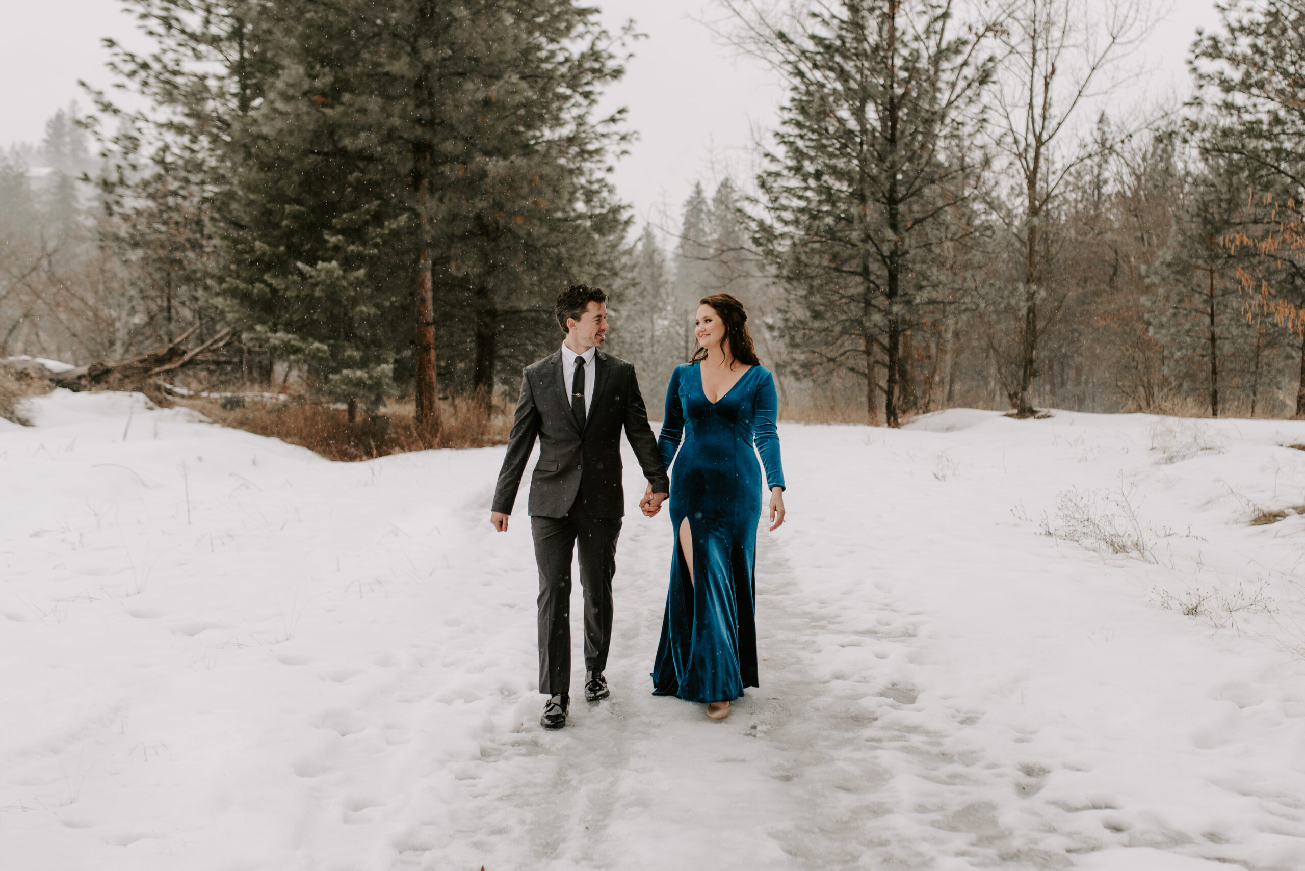 A future bride and groom walking in the snow together for their engagement photos.