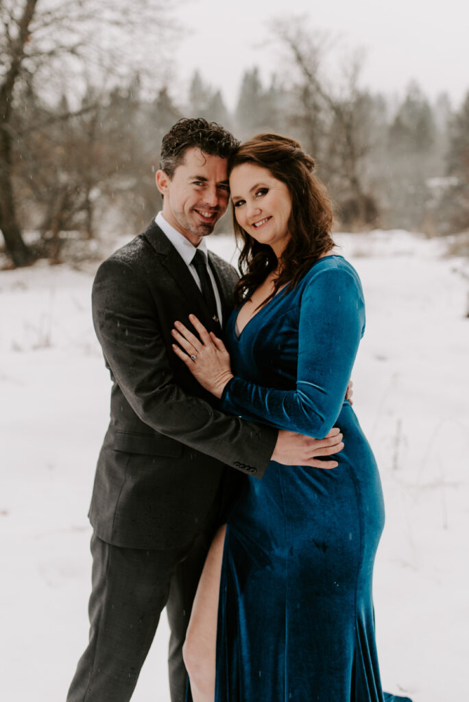 An engaged couple hugging and smiling at the cameras during their engagement photos in the snow.