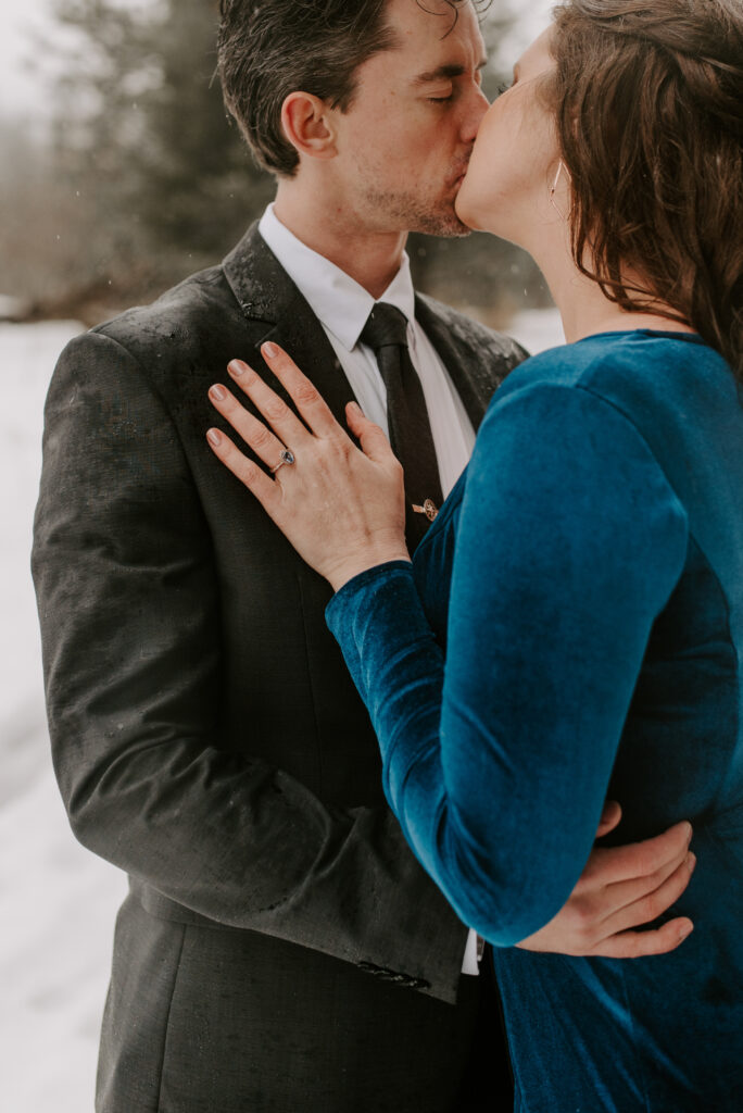 An engaged couple kissing while it's snowing outside.