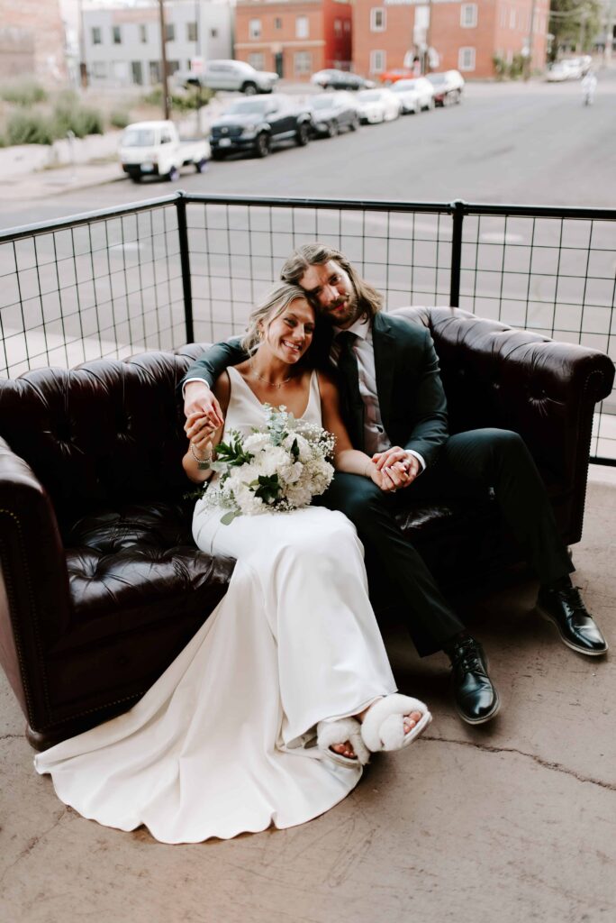 Bride and Groom sitting on a leather couch and smiling at the camera.